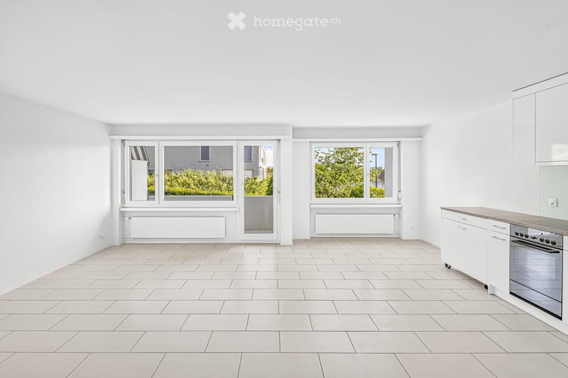 Empty room with white walls, tiled floor, large windows, and a small kitchen area with cabinets and an oven.