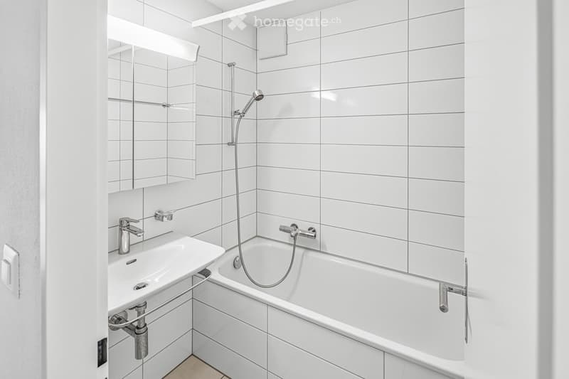 Modern bathroom with white tiled walls and floor, featuring a bathtub with a handheld showerhead, a sink with a modern faucet, and built-in storage shelves above the sink.