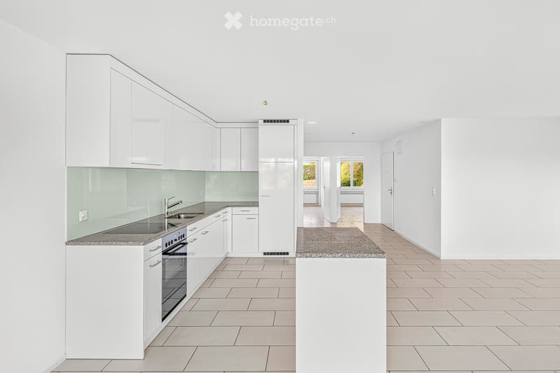 Modern, minimalist kitchen with white cabinets, granite countertops, and tiled flooring. The space is open and uncluttered, featuring built-in appliances and ample natural light from nearby windows.