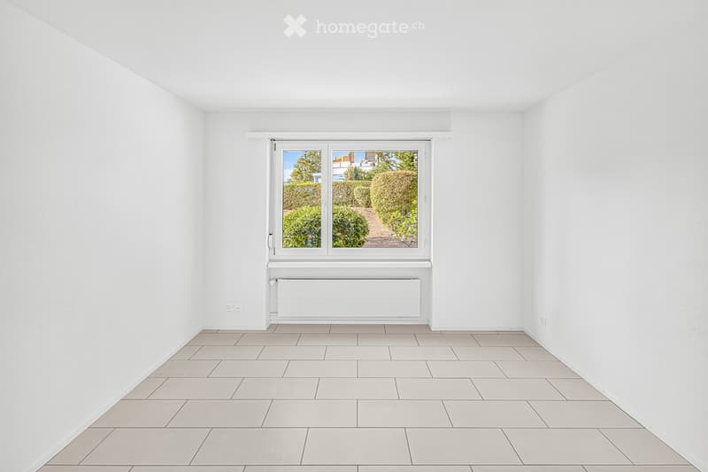 Empty room with white walls, tiled floor, and a large window showing a garden view. No furniture or equipment present.