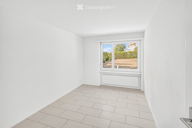 Empty room with white walls, tiled floor, and a large window showing a view of greenery and a building outside.