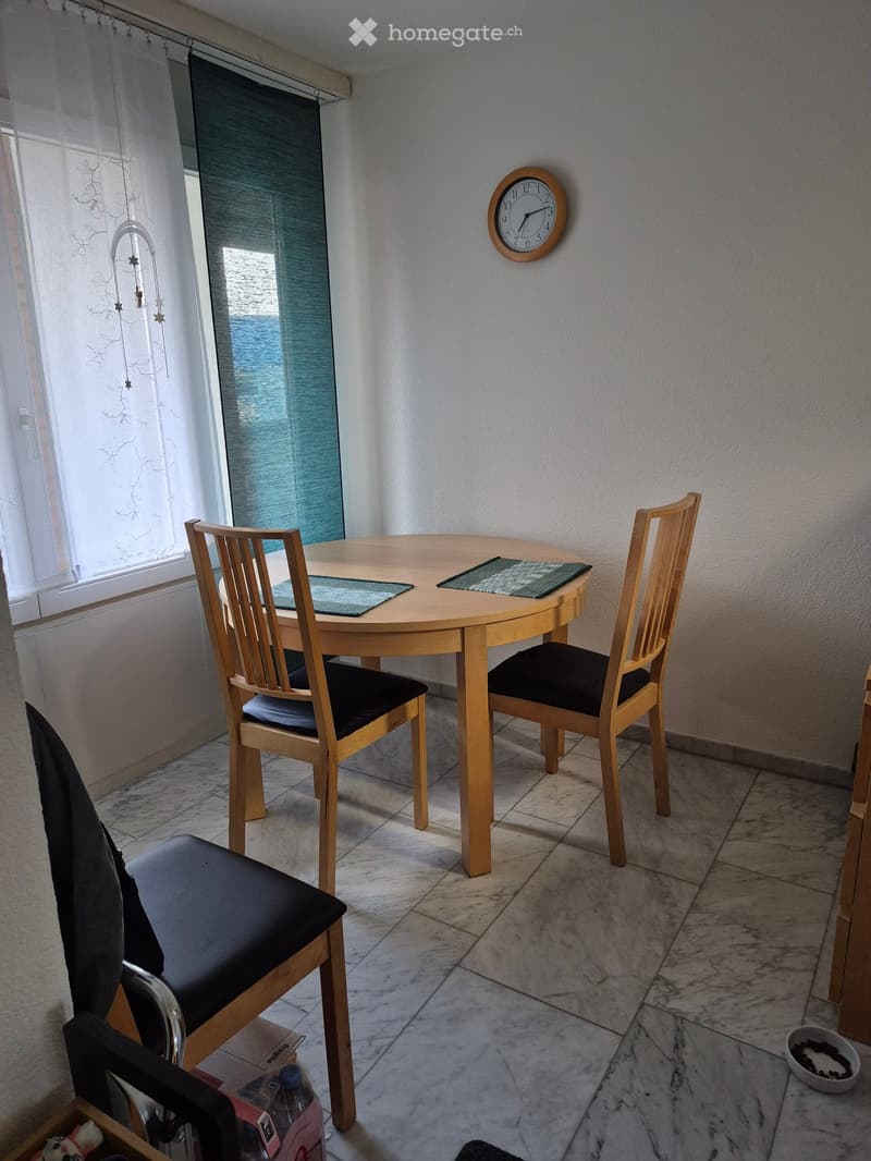 Dining area with a round wooden table, four chairs with black cushions, a wall clock, and placemats on the table.