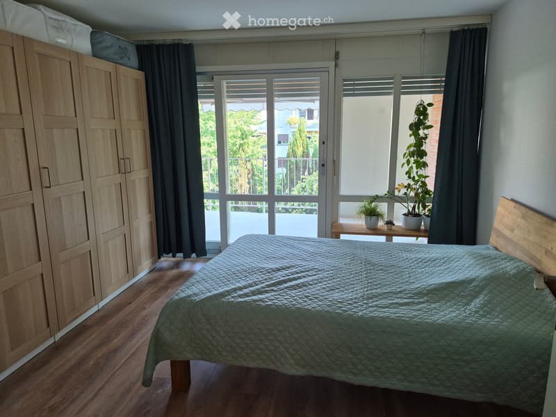 Bright bedroom with wooden floor, large window with balcony access, bed with green quilt, wardrobe, small table with potted plants.