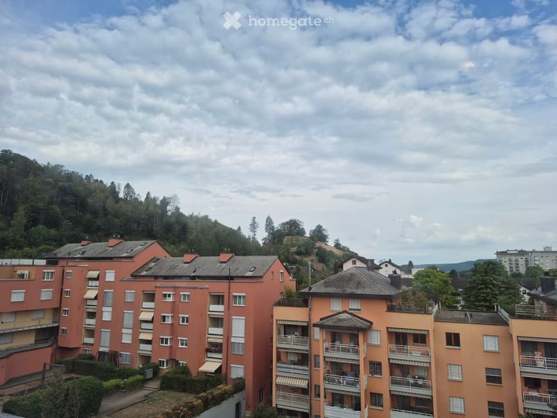 Multi-story residential building with balconies and a view of a forested hillside in the background.