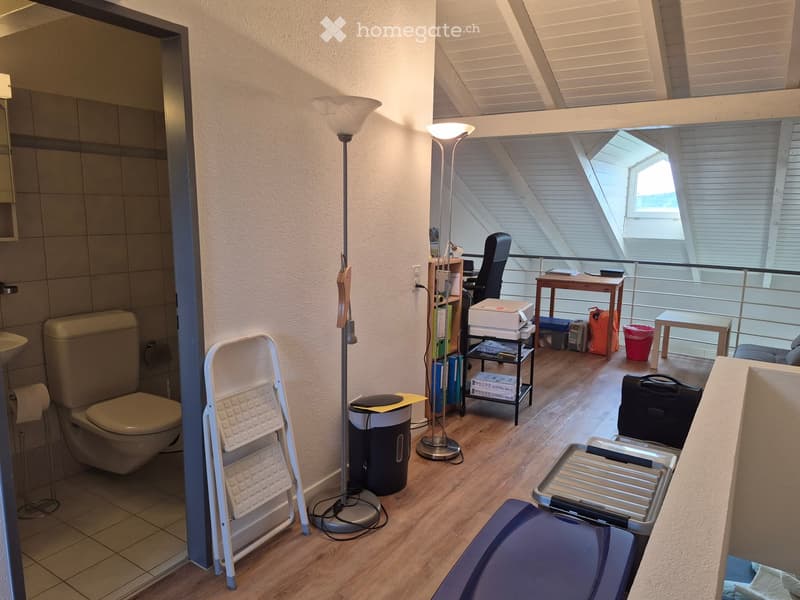 The image shows a furnished room with a toilet, sink, and a small workspace area with a chair, table, and laptop. There are several stools and a trash bin visible.