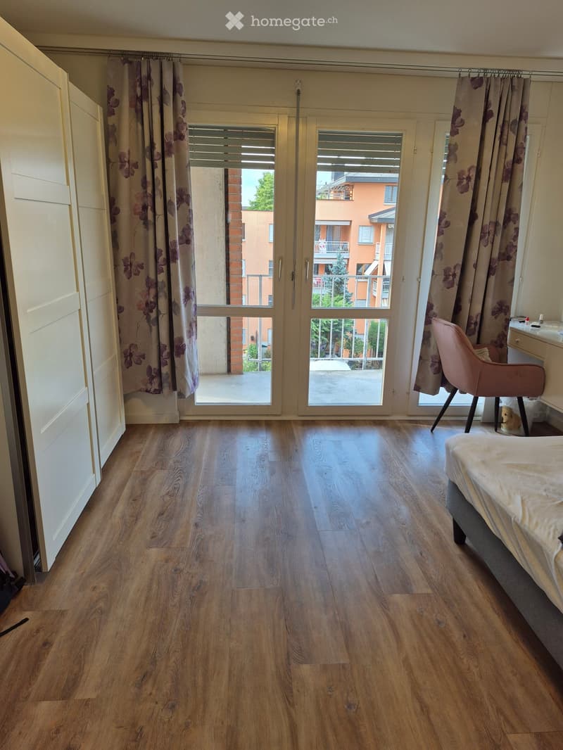 Bright bedroom with wooden flooring, large windows with floral curtains, and a view of the balcony and surrounding buildings.