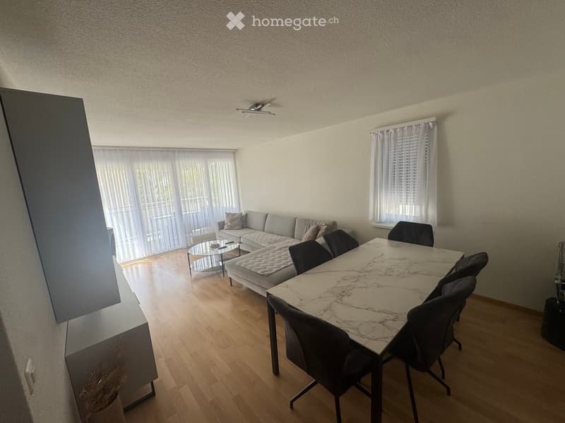 Modern living and dining area with a grey sectional sofa, coffee table, marble dining table, and chairs. Large windows with sheer curtains and wooden flooring.