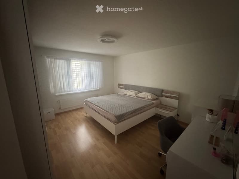 Well-lit bedroom with wooden flooring, a bed with a headboard, a nightstand, a desk with a chair, and a mirror on the wall.