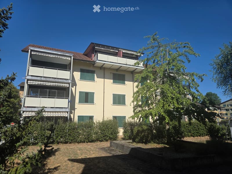 A three-story residential building with beige walls, green shutters, and multiple balconies. The building is surrounded by trees and shrubs, with a raised ground floor visible in the foreground.