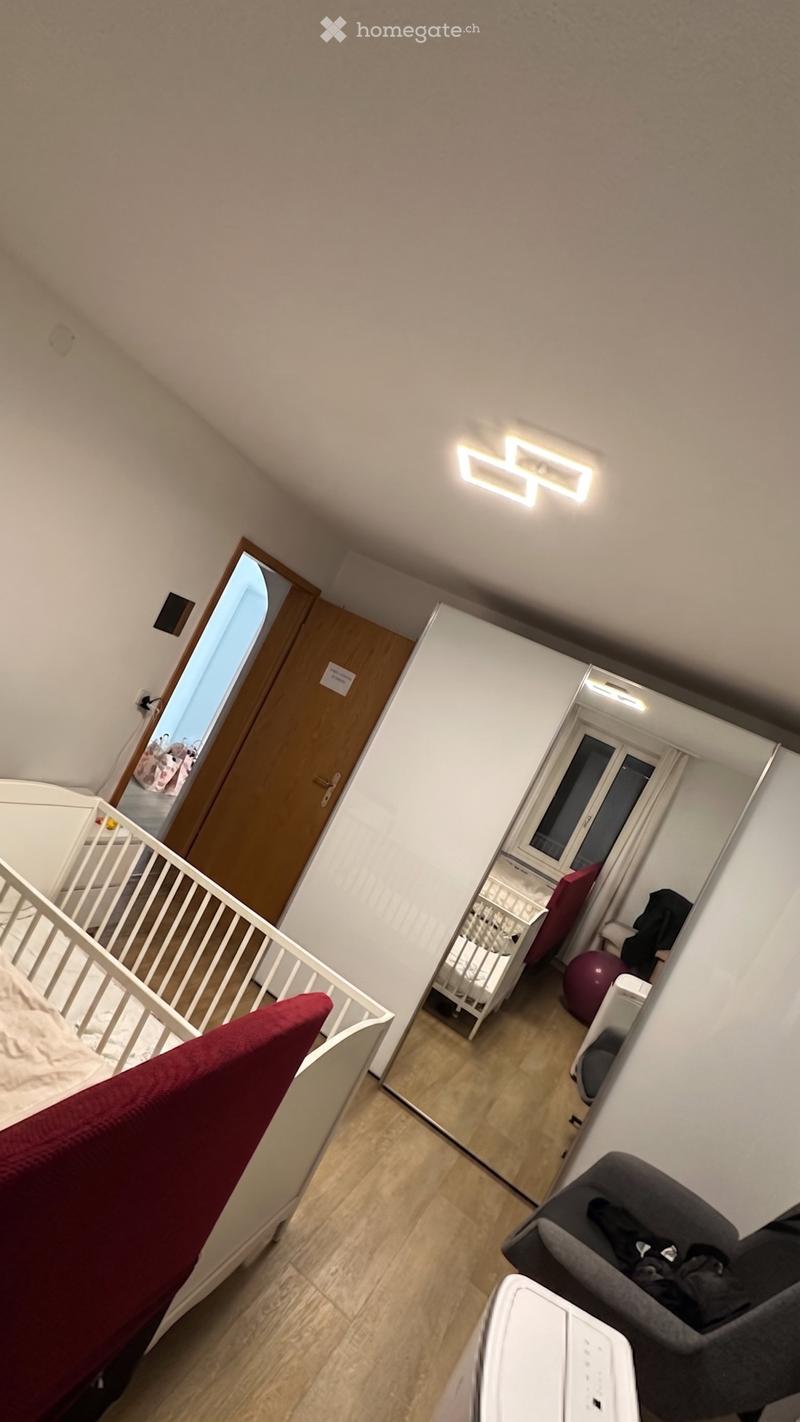 A small, well-lit room with wooden flooring, a baby crib, a baby chair, a small table, and a ball in the corner.