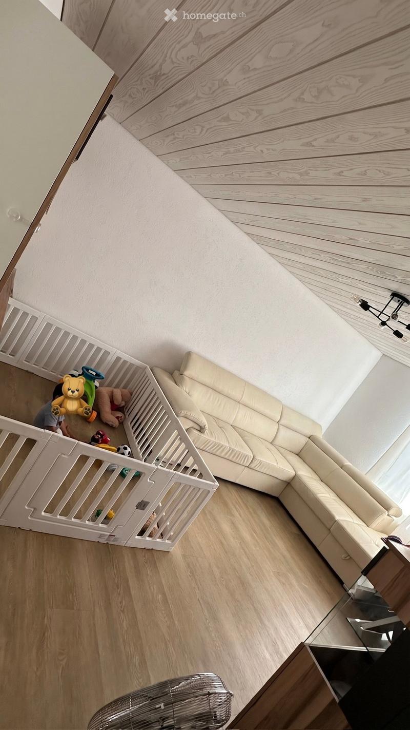 A living room with a white baby gate, a beige sectional sofa, a glass coffee table, a fan, and toys inside the gate.