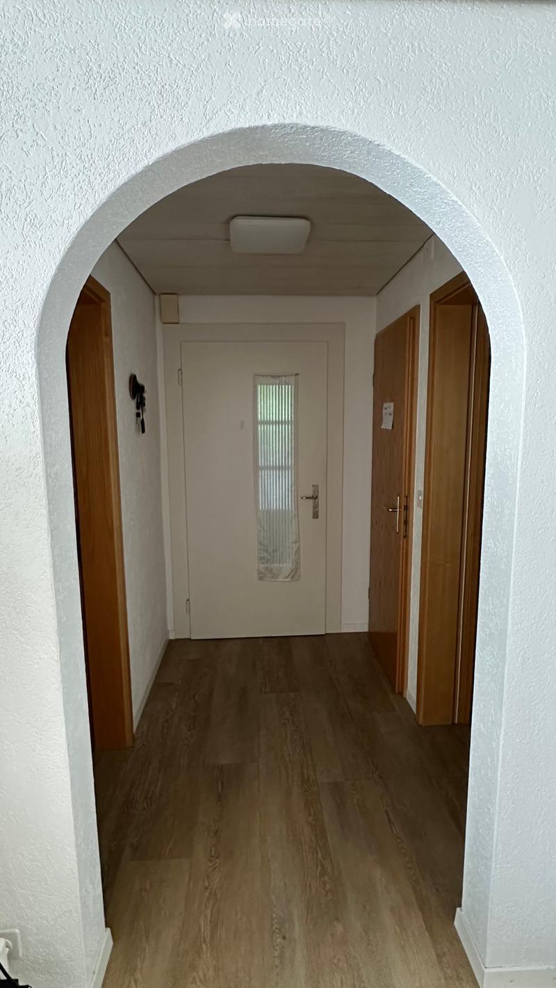 A clean, well-lit hallway with wooden flooring, white walls, and several wooden doors on both sides. An arched ceiling adds architectural interest.