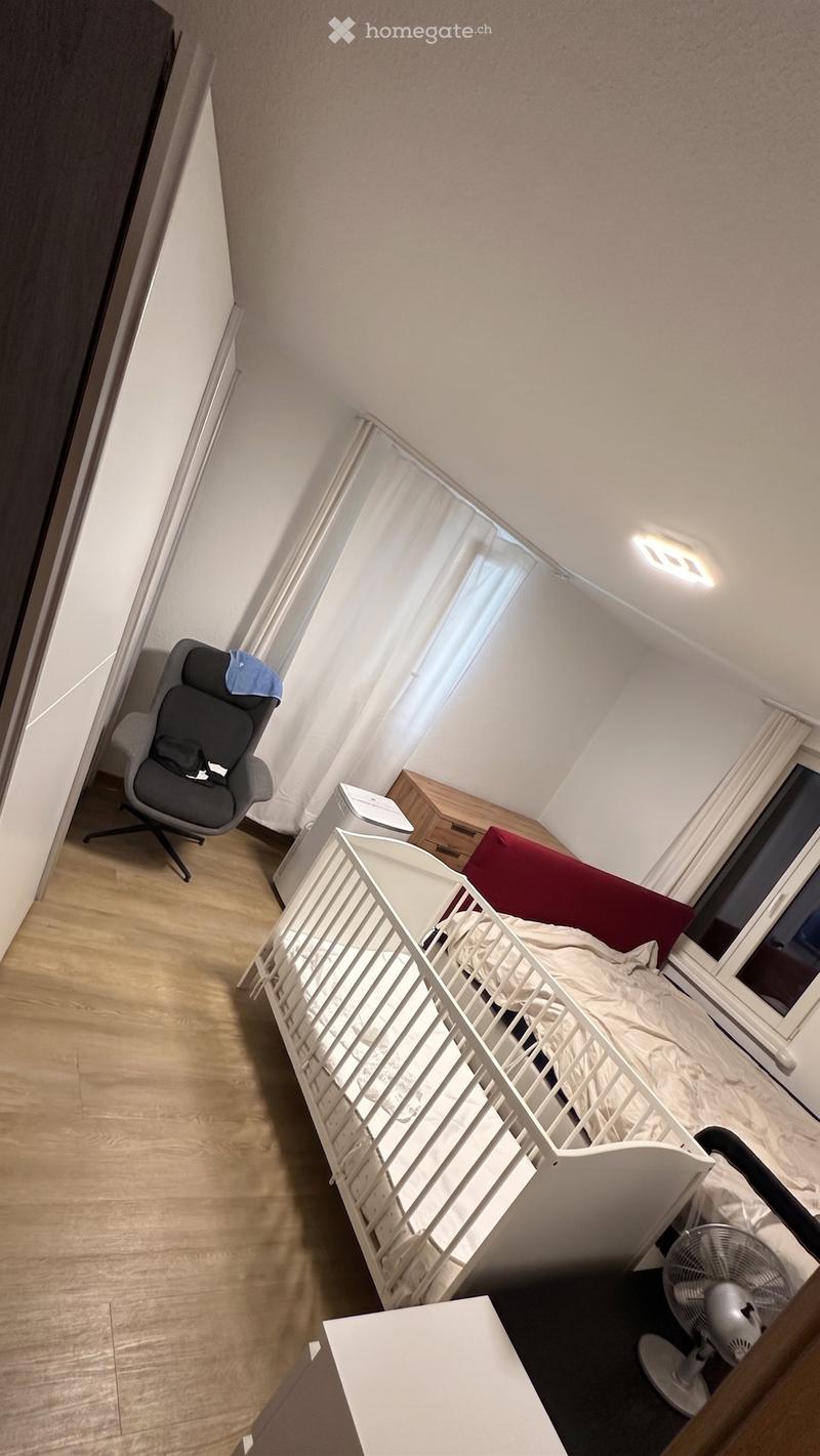 A small bedroom with a baby crib, a chair, a fan, and a nightstand. The room has wooden flooring and large windows with curtains.
