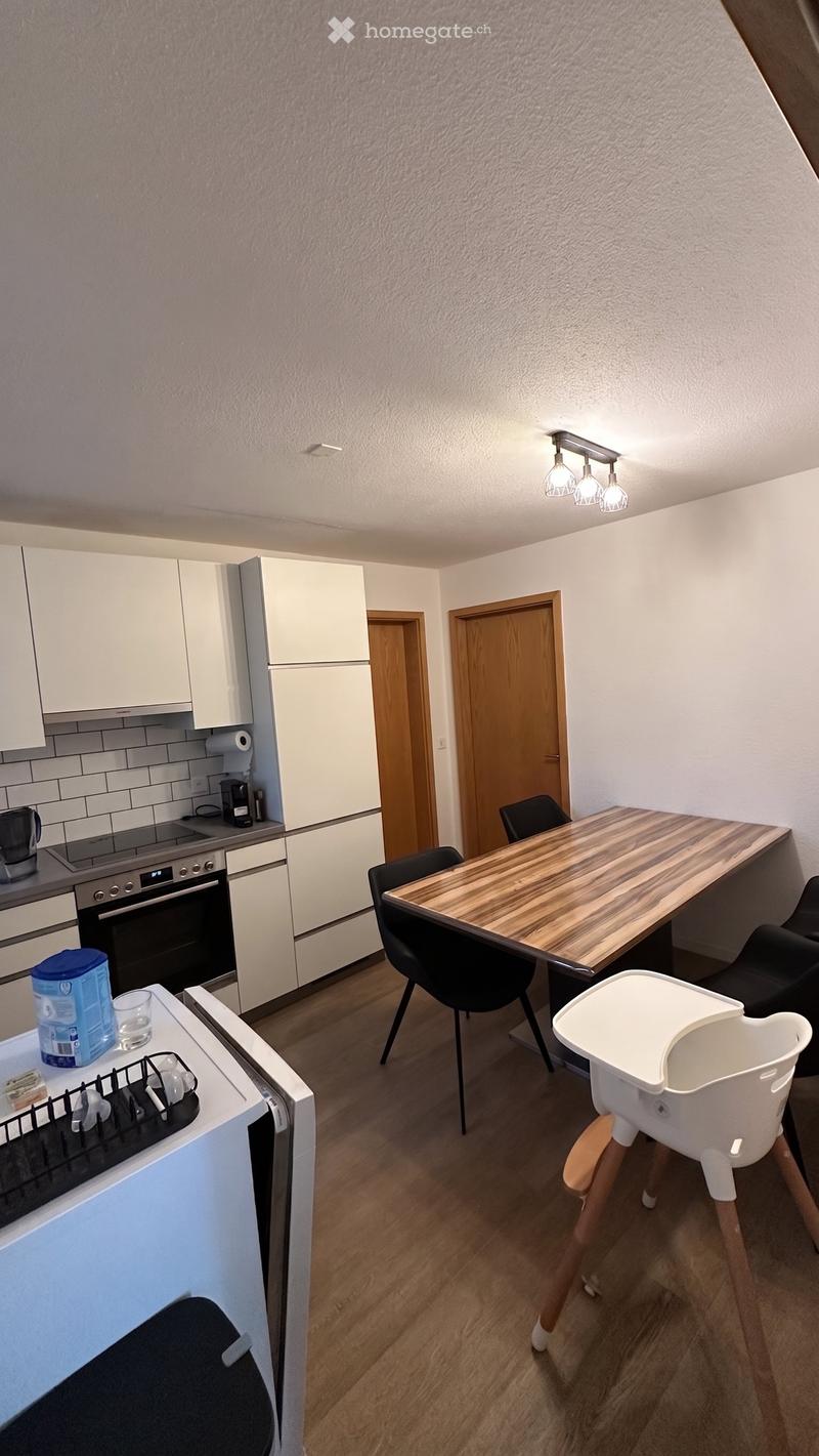A modern kitchen and dining area with white cabinets, a wooden dining table, black chairs, a baby chair, and various appliances including an oven and fridge.