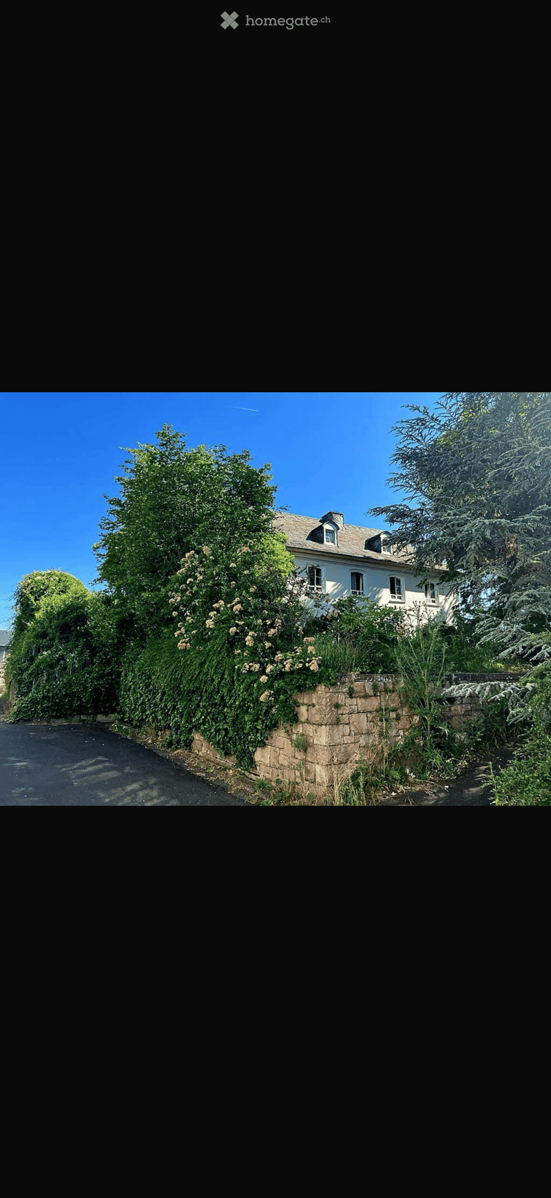 A two-story white house with multiple windows, surrounded by lush greenery and a stone wall. The house features a gray roof and is partially obscured by trees and flowering shrubs.