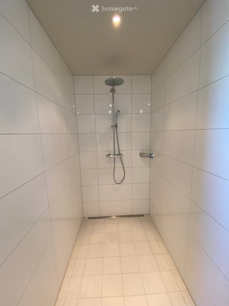 Modern shower stall with white tiled walls and floor, featuring a handheld showerhead and a built-in towel rack.