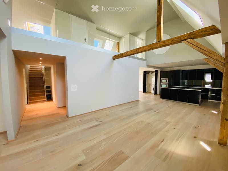 Open-plan space with light wooden flooring, white walls, and exposed wooden beams. Features a staircase leading to an upper level, a modern kitchen area with black cabinets and stainless steel appliances, and a balcony area with large windows.