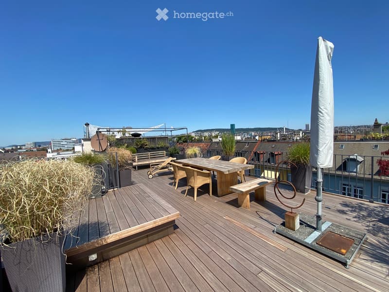 Wooden deck terrace with a table, chairs, bench, umbrella, and potted plants, overlooking a cityscape.