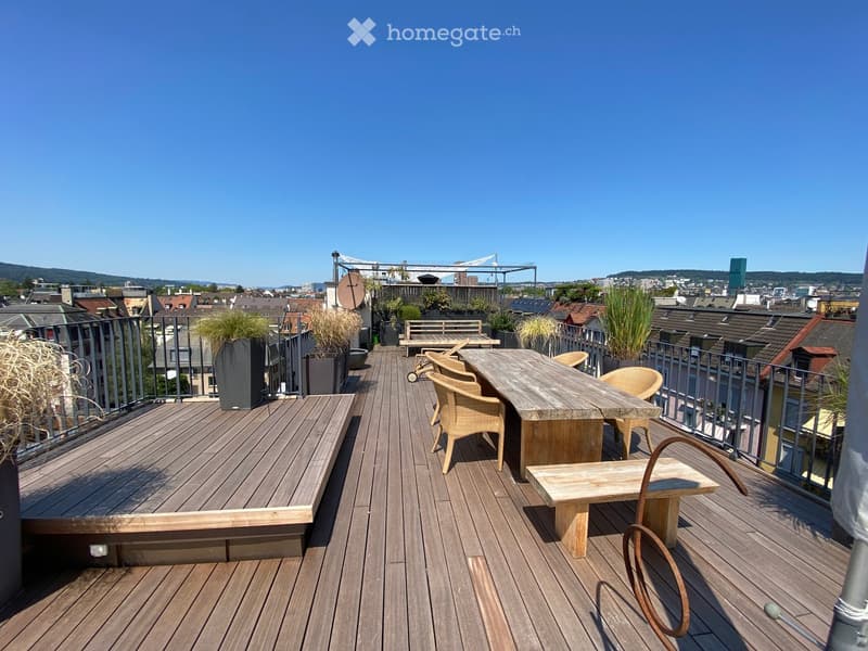 Wooden deck terrace with a table, chairs, bench, and potted plants, overlooking a cityscape.
