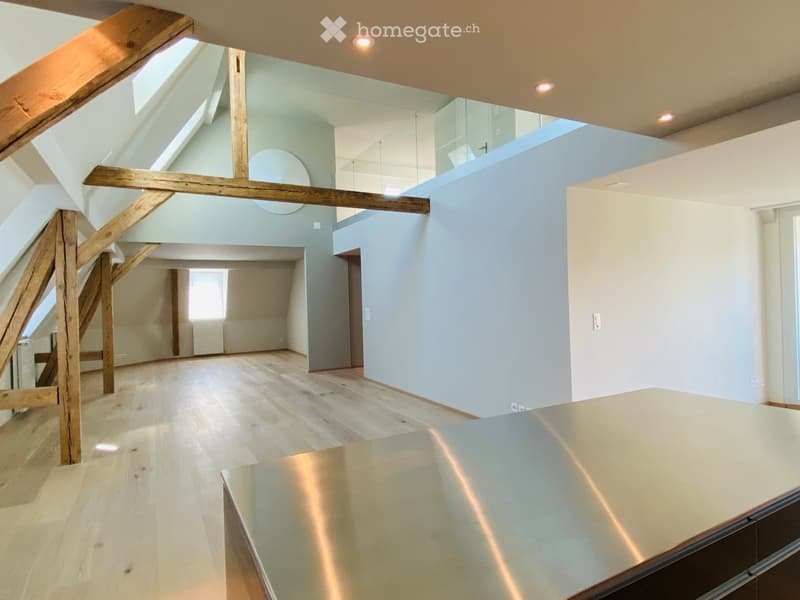 Open, airy space with wooden floors, exposed wooden beams, and large windows allowing natural light. The room features a modern, minimalist design with a glass railing and a sleek, metallic countertop in the foreground.