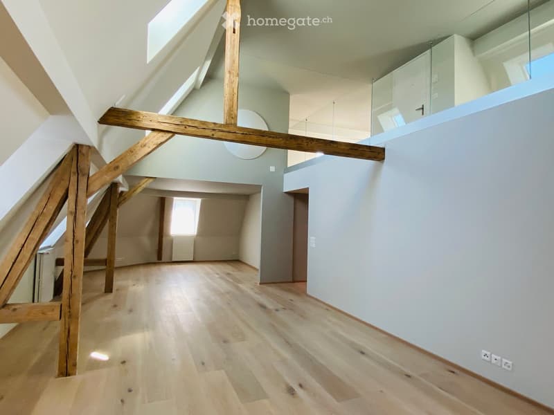 Bright, open hallway with wooden flooring, exposed wooden beams, and a staircase leading upwards. Natural light streams in from skylights and windows, creating a spacious and airy atmosphere.