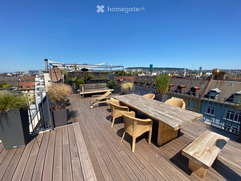 Wooden deck with a table, chairs, a bench, and a lounge chair, surrounded by potted plants and offering a cityscape view.