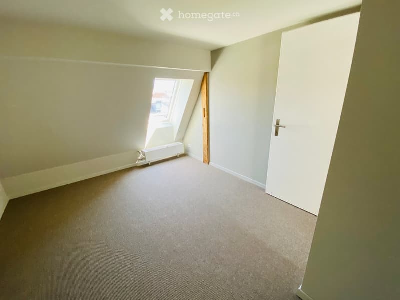 Empty attic room with sloped ceilings, carpeted floor, white walls, a small window, and a white door.