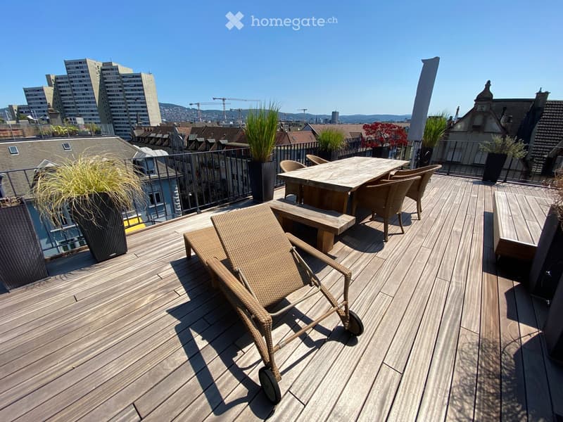 Wooden deck with a table, chairs, a bench, and a lounge chair, surrounded by potted plants and a cityscape view.