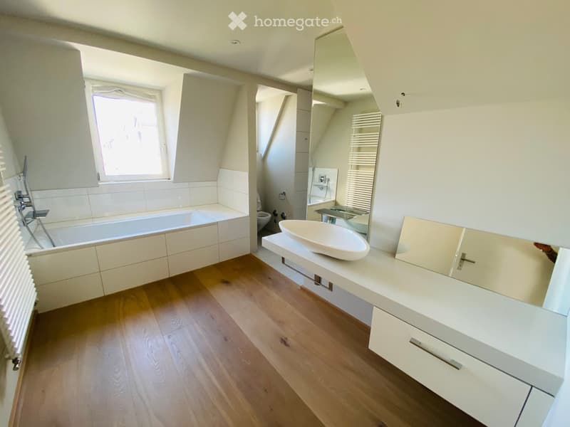 Modern bathroom with a bathtub, sink, mirror, radiator, and toilet. The space features light-colored tiles, wooden flooring, and ample natural light from a window.