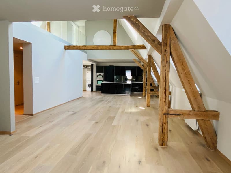 Empty hallway with wooden flooring, light blue walls, and exposed wooden beams. The hallway leads to a kitchen area with dark cabinets and stainless steel appliances visible in the background.