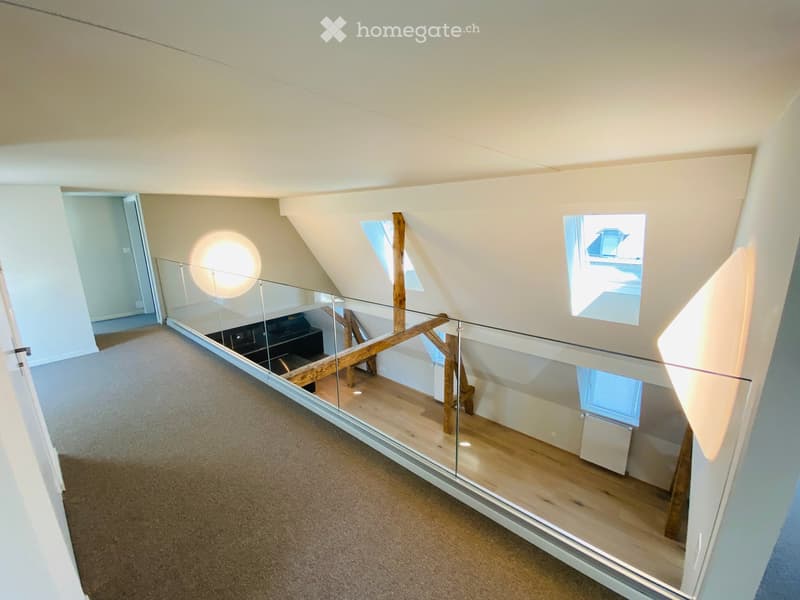 Spacious, open corridor with a glass railing leading to a lower level, wooden beams, and skylights providing natural light.