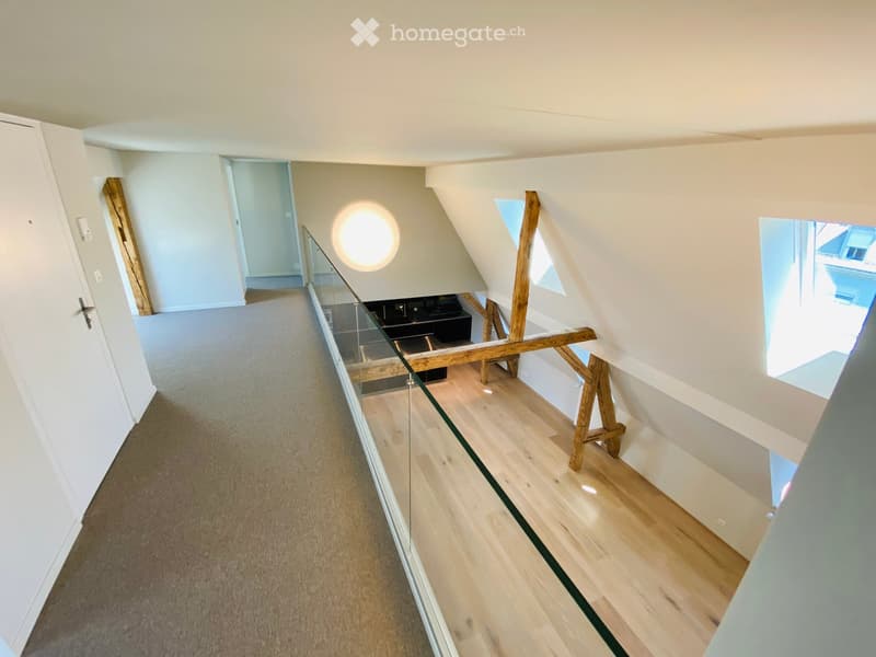 Modern hallway with wooden flooring, glass railing, and exposed wooden beams. Features a round light fixture and open doorways leading to other rooms.
