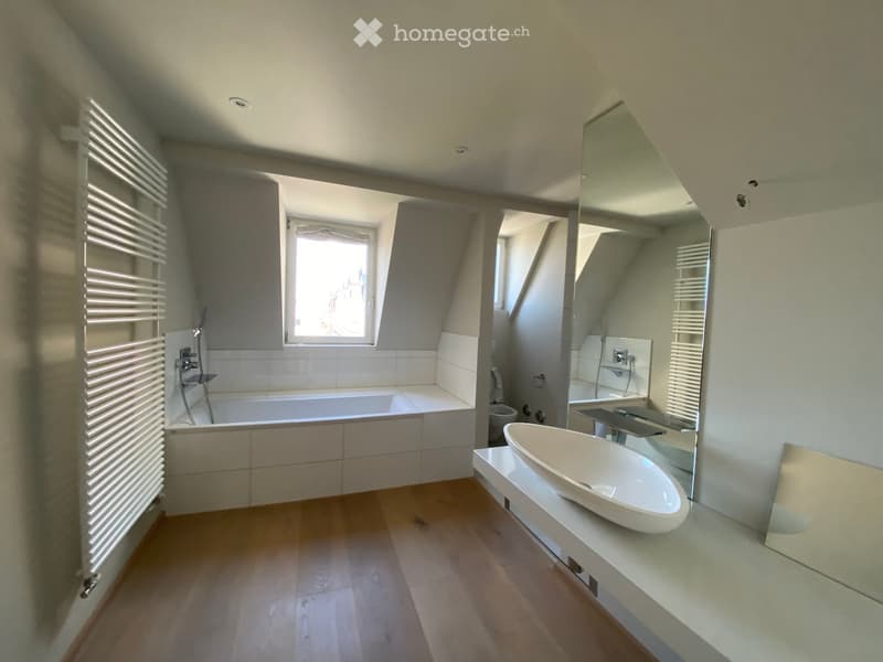 Modern bathroom with a bathtub, sink, toilet, radiator, and large mirror. The space features wooden flooring and a window letting in natural light.