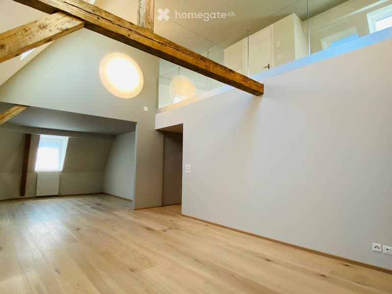 Bright, open loft-style room with wooden flooring, exposed wooden beams, and large windows allowing natural light. The space features a modern glass railing leading to an upper level.