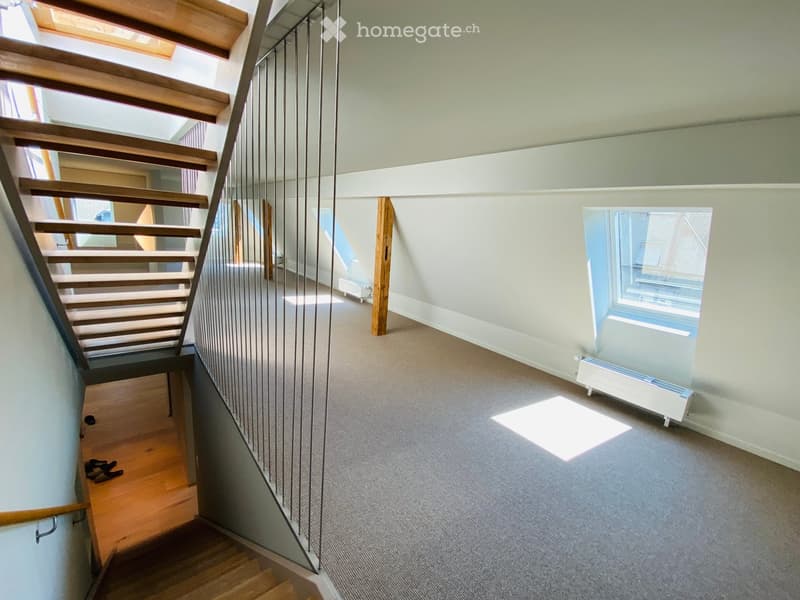 Modern hallway with wooden stairs, metal railing, carpeted floor, and large window letting in natural light.