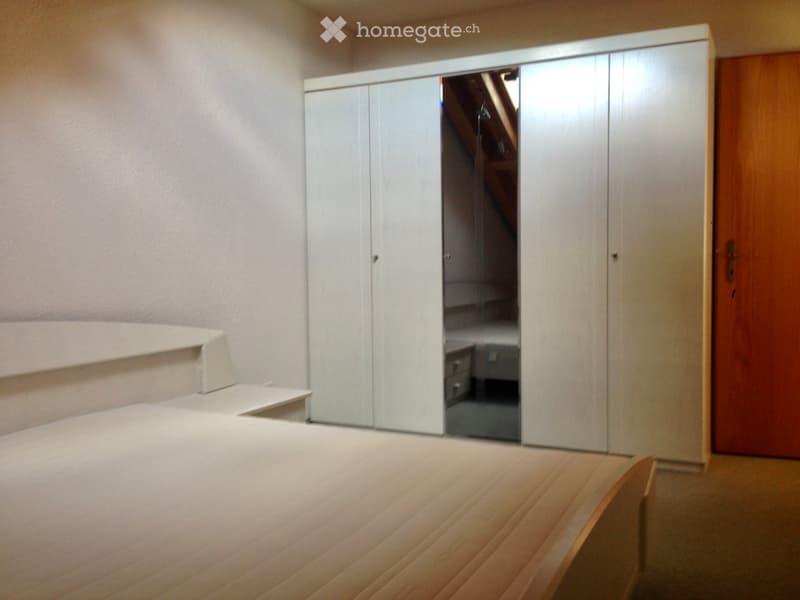 A small, furnished room with a white wardrobe, a wooden chair, and a staircase leading upwards.