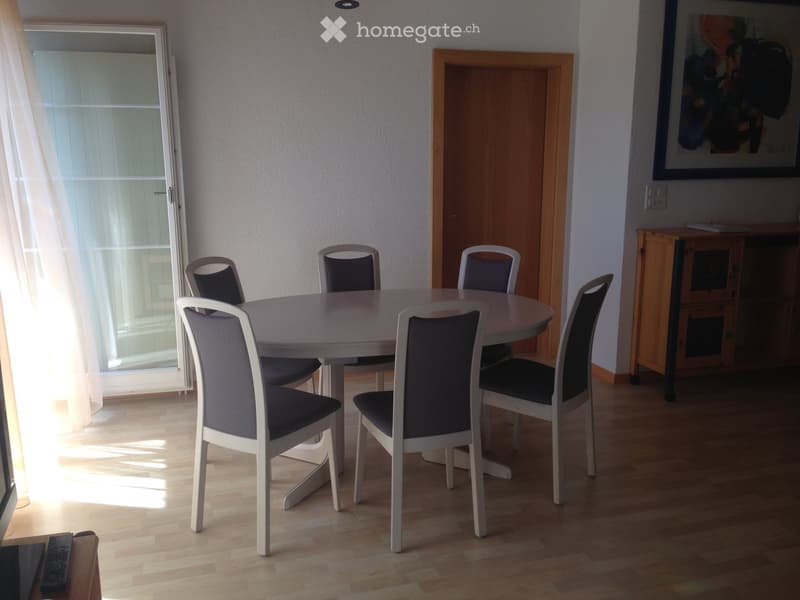 Dining room with a round white table surrounded by six chairs, a wooden cabinet, and a large window with white curtains.