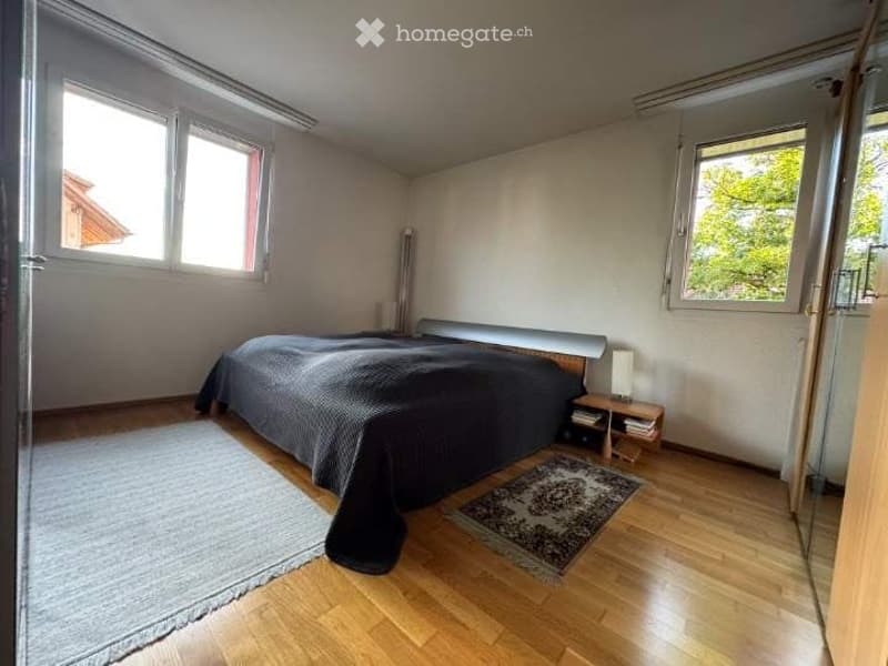 Spacious bedroom with wooden flooring, large windows, a neatly made bed with dark bedding, a nightstand with a lamp, and two rugs on the floor.