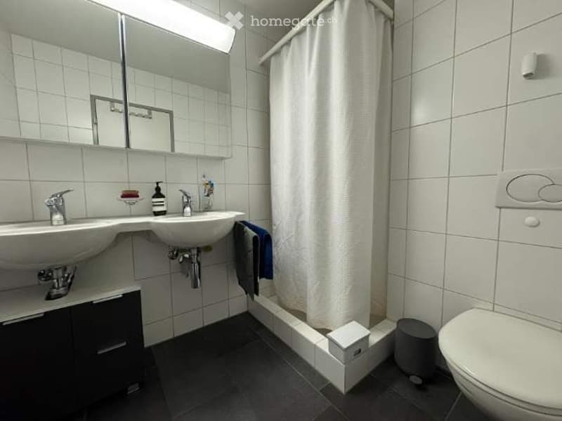 Modern bathroom with two sinks, a large mirror, a shower area with a curtain, and a toilet. The walls are tiled in white, and the floor is dark grey.