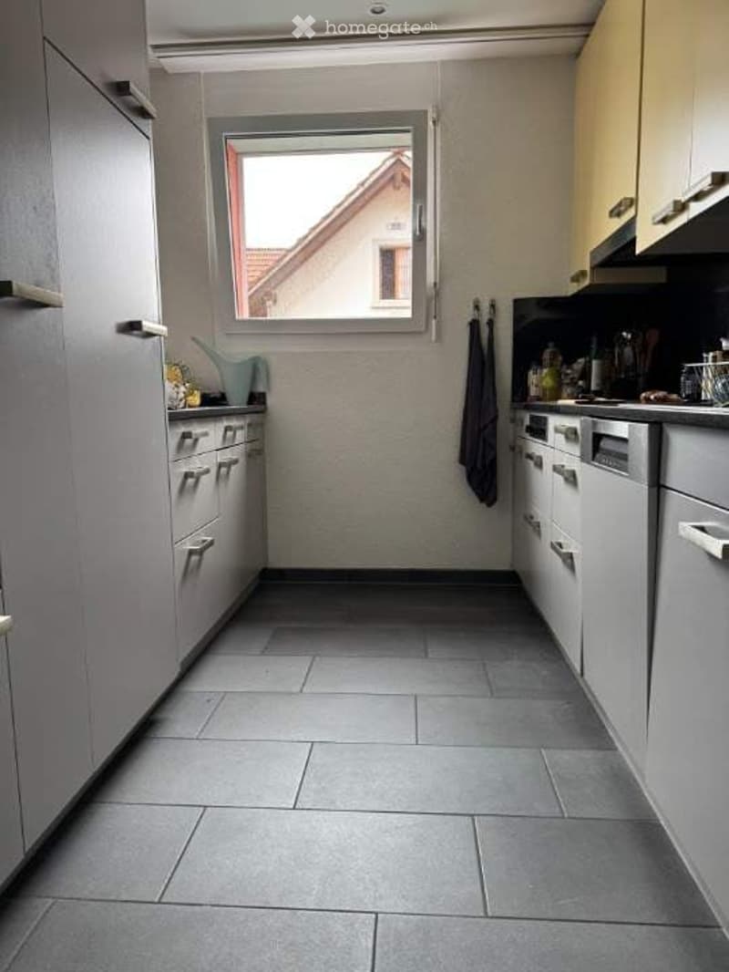 Modern kitchen with grey tiled floor, white cabinets, and stainless steel appliances including a dishwasher and oven. A window offers a view of a neighboring building.