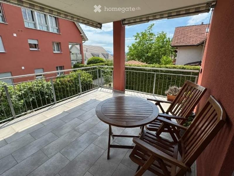 Balcony with tiled floor, wooden table, and chairs, overlooking a residential area with greenery and buildings.