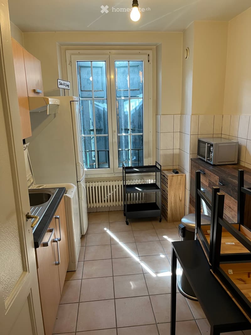 Compact kitchen with tiled floor, white refrigerator, microwave, sink, and a small cart. Natural light streams in from a windowed door.