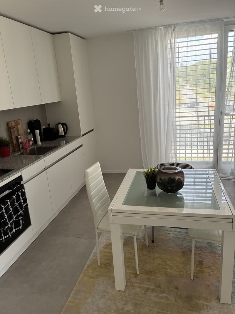 Modern kitchen with white cabinets, a glass dining table with a white chair, and various appliances including an oven, sink, and coffee maker. The room features large windows with white curtains and a small potted plant on the counter.