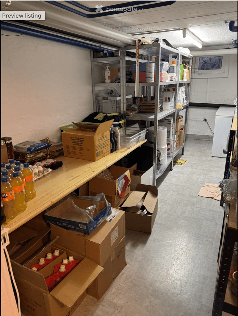 Storage area with metal shelving units, wooden shelves, and various boxes and supplies, including bottled drinks and cleaning products.
