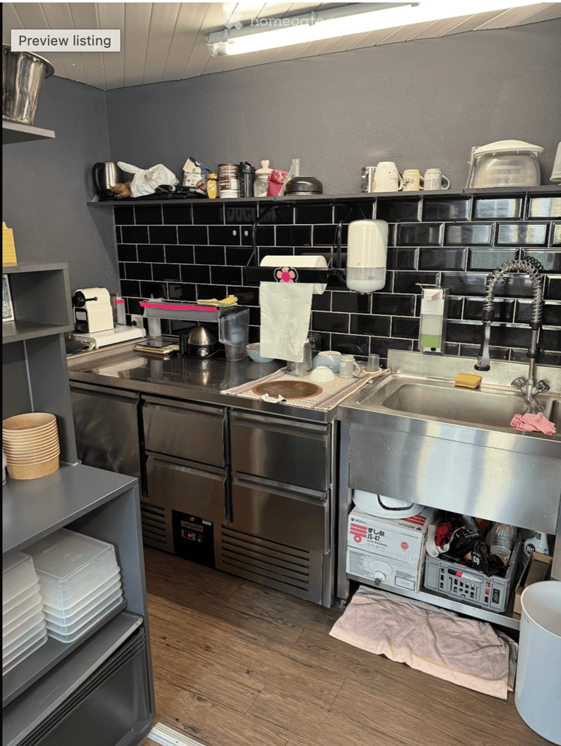 The kitchen features stainless steel appliances, including a sink, dishwasher, oven, and refrigerator. There are wooden shelves with various kitchen items, including cups, bowls, and containers. The floor is wooden, and the walls are dark with black tiles