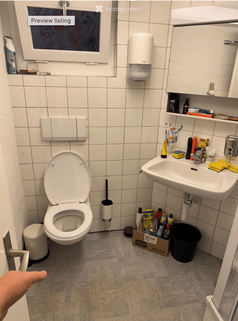 A small bathroom with white tiled walls and a grey tiled floor. It features a toilet, a sink with a faucet, a mirror, and various toiletries including toothpaste, soap, and cleaning supplies. A hand sanitizer dispenser is mounted on the wall, and a trash
