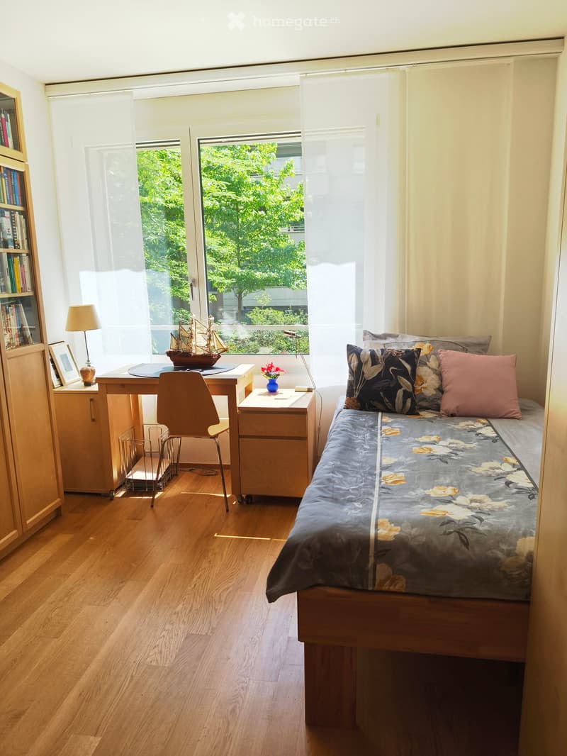 Cozy bedroom with wooden flooring, a single bed with floral bedding, a desk with a model ship and lamp, a nightstand with a vase of flowers, and a bookcase with books and a picture frame.