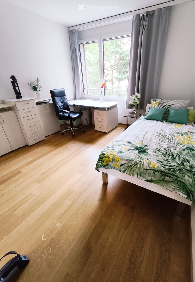 Bright bedroom with wooden flooring, large window with view, bed with green and yellow bedding, desk with chair, side table, and decorative vases.