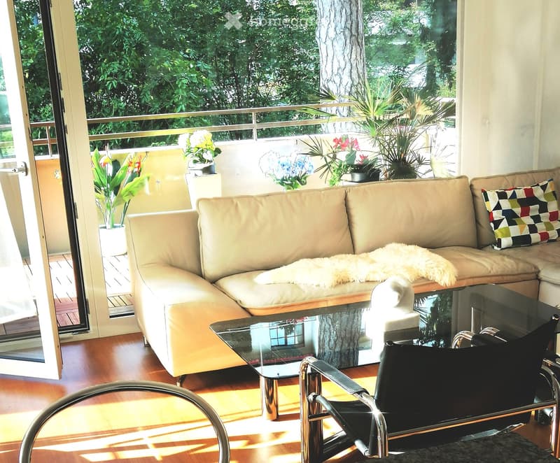 Open living room with large glass doors to balcony, beige couch with pillow, glass coffee table, wooden floor, balcony with view of greenery