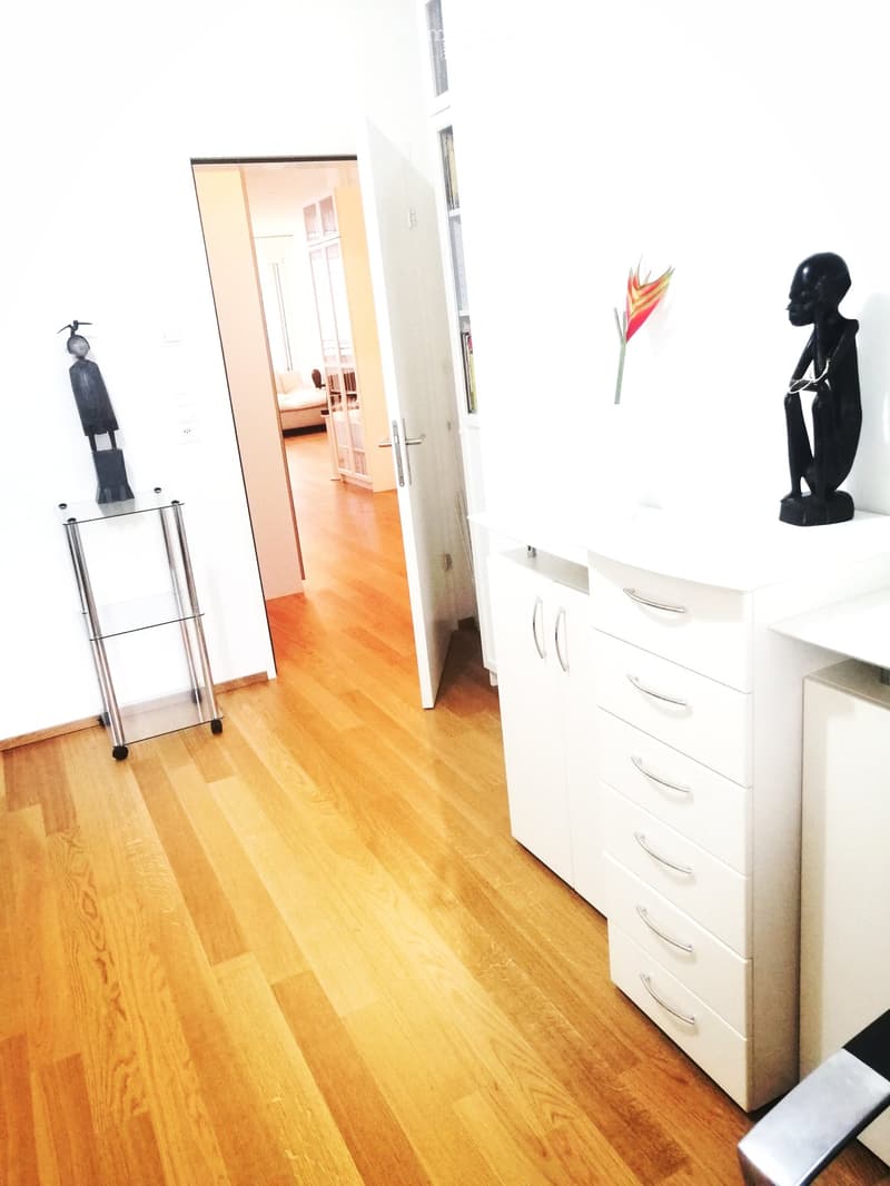 Open layout with wooden floor, white walls, two black statues, open door leading to another room, white cabinets, flower on the top of the cabinets